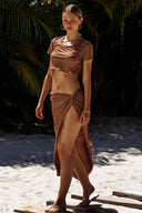 Woman in a brown outfit standing on a sandy beach with greenery in the background