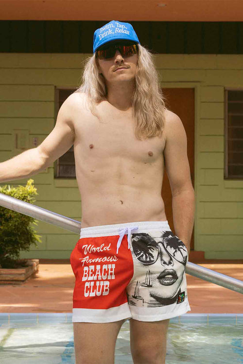 Man wearing a blue cap and red and white swim shorts with a graphic design, standing by a pool.