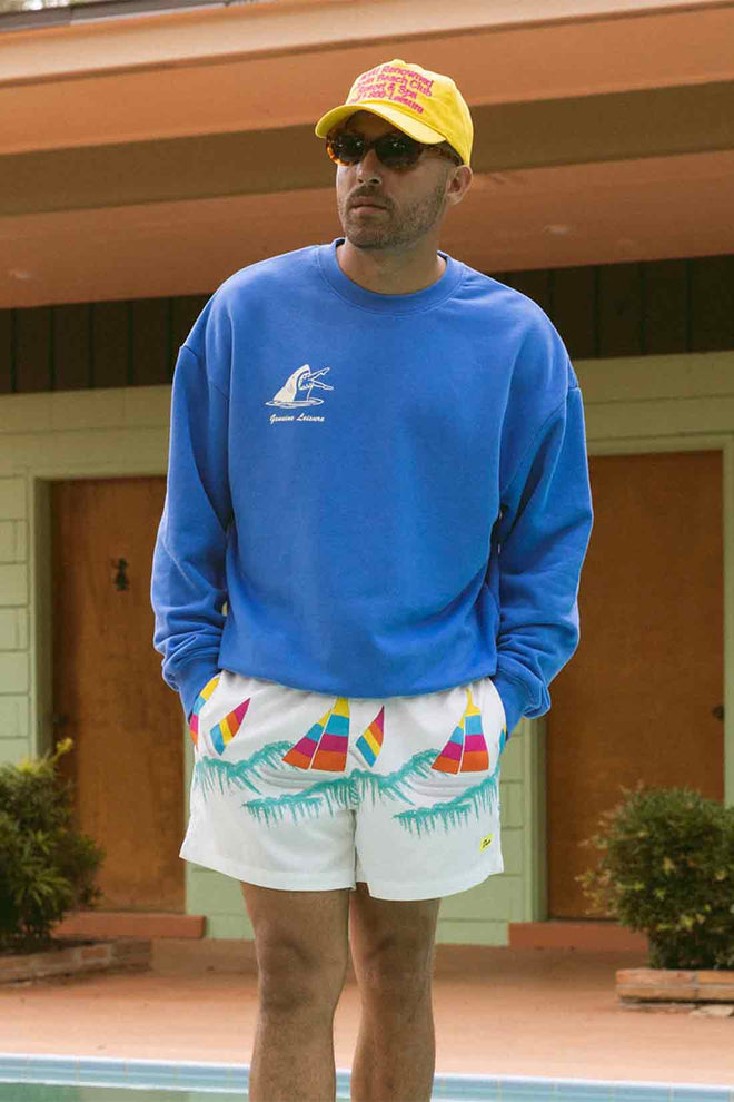 Man wearing a blue sweatshirt, yellow cap, and colorful shorts standing by a pool.