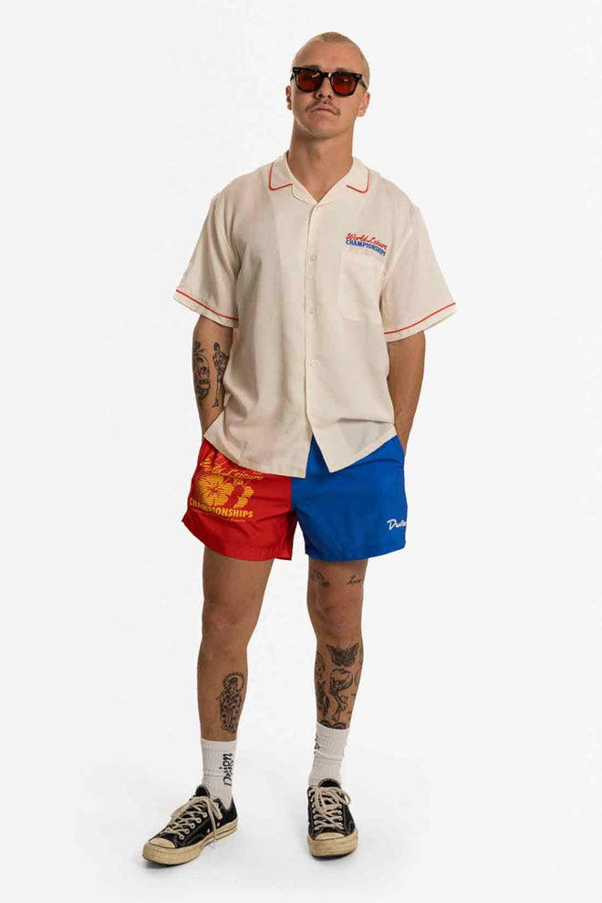 Man wearing a beige baseball jersey, red and blue shorts, and sunglasses on a white background